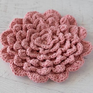 May include: A pink crocheted flower with a layered design. The flower is made with a soft yarn and has a delicate texture.