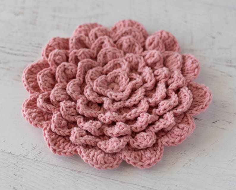 May include: A pink crocheted flower with a layered design. The flower is made with a soft yarn and has a delicate texture.