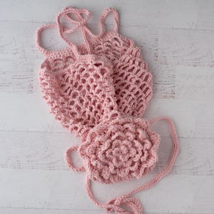 May include: A light pink crochet bag featuring an open mesh net construction and a large, dimensional floral appliqu&eacute; attached to the front. The bag includes two thin, braided crochet straps for carrying and is crafted from soft yarn.