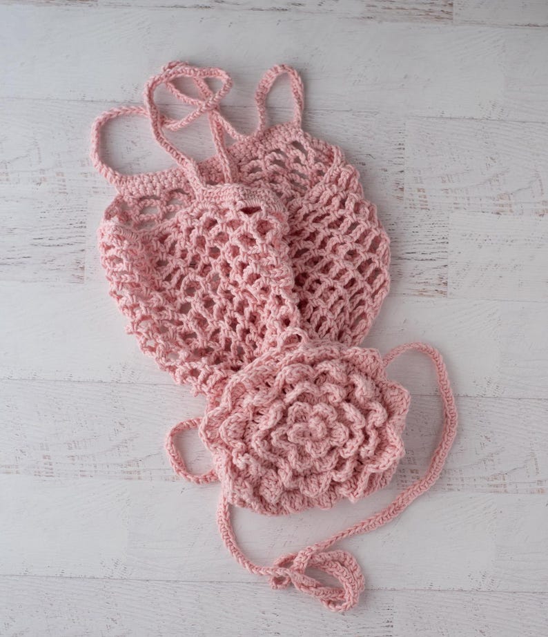 May include: A light pink crochet bag featuring an open mesh net construction and a large, dimensional floral appliqu&eacute; attached to the front. The bag includes two thin, braided crochet straps for carrying and is crafted from soft yarn.
