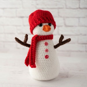 May include: A white crocheted snowman with a red knit hat and scarf. The snowman has brown twig arms and red and white buttons.