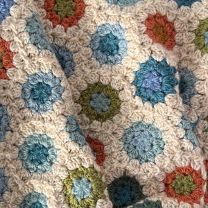 May include: Close-up of a handmade crochet blanket. The blanket features a repeating pattern of colorful floral motifs in shades of blue, green, orange, and cream. The texture of the yarn is visible.