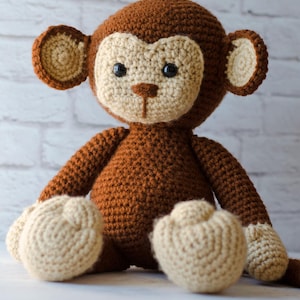 May include: A brown crocheted monkey toy with beige ears, hands, and feet. The monkey has black eyes and a beige nose.