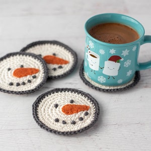 K&ouml;nnte beinhalten: Vier geh&auml;kelte Schneemann-Untersetzer mit orangenen Nasen und schwarzen Augen und eine blaue Tasse mit einem wei&szlig;en Design aus zwei Marshmallows, die sich die H&auml;nde halten. Die Tasse ist mit hei&szlig;er Schokolade gef&uuml;llt.