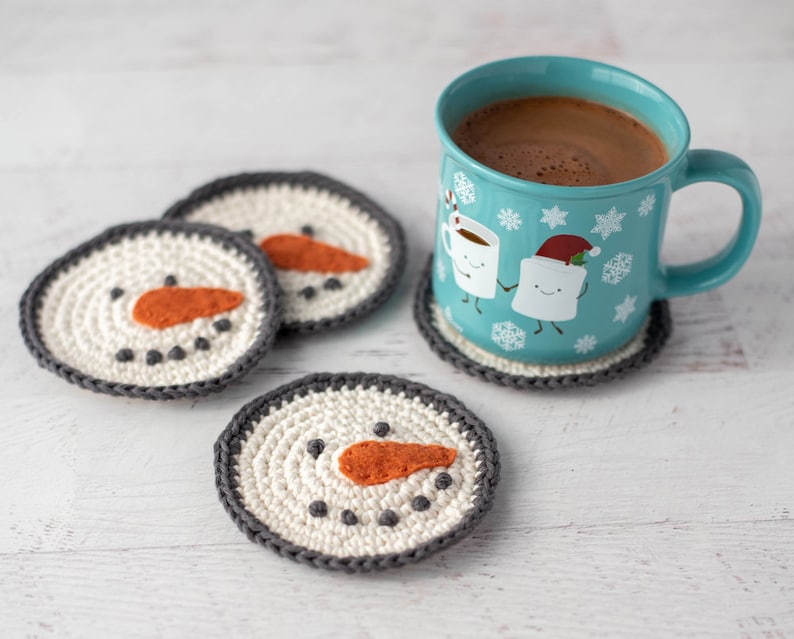 K&ouml;nnte beinhalten: Vier geh&auml;kelte Schneemann-Untersetzer mit orangenen Nasen und schwarzen Augen und eine blaue Tasse mit einem wei&szlig;en Design aus zwei Marshmallows, die sich die H&auml;nde halten. Die Tasse ist mit hei&szlig;er Schokolade gef&uuml;llt.