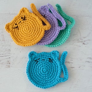May include: Set of five crocheted cat-shaped coasters in yellow, purple, green, and blue. The coasters have a simple design with a cat face and a tail.