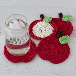 May include: Three round, red crochet apple coasters and one crochet apple slice coaster with a cream-colored center. Each coaster features a small brown stem and a light green leaf. A clear glass jar filled with water and ice cubes sits on one coaster.