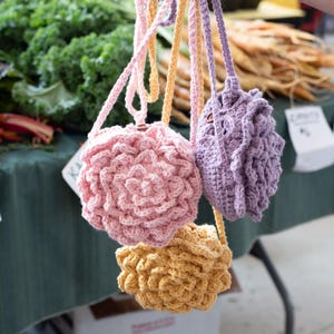 May include: Three small, round, crocheted bags in light pink, lavender, and yellow. Each bag features a textured, layered floral pattern on the front and includes a matching crocheted strap for carrying. The bags are held together by their straps.