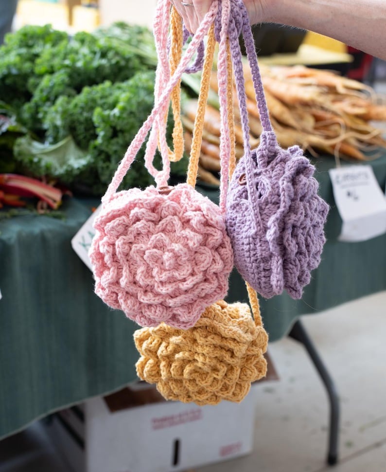 May include: Three small, round, crocheted bags in light pink, lavender, and yellow. Each bag features a textured, layered floral pattern on the front and includes a matching crocheted strap for carrying. The bags are held together by their straps.