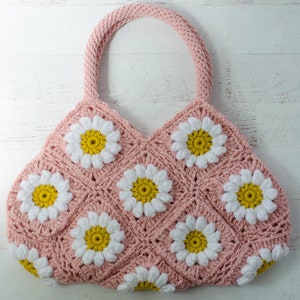 May include: A pink crocheted tote bag with white and yellow daisy flowers. The bag has a single handle.
