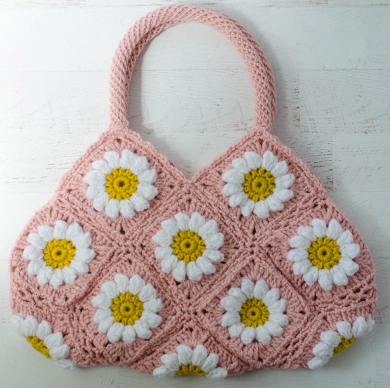 May include: A pink crocheted tote bag with white and yellow daisy flowers. The bag has a single handle.