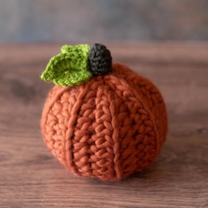 May include: A crocheted pumpkin with a green stem and a black centre. The pumpkin is a light orange colour and is made with a chunky yarn.