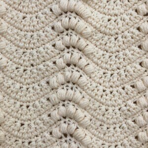 May include: A close-up of a crocheted blanket with a textured pattern. The blanket is made of a cream-colored yarn and has a repeating pattern of small, raised bumps.