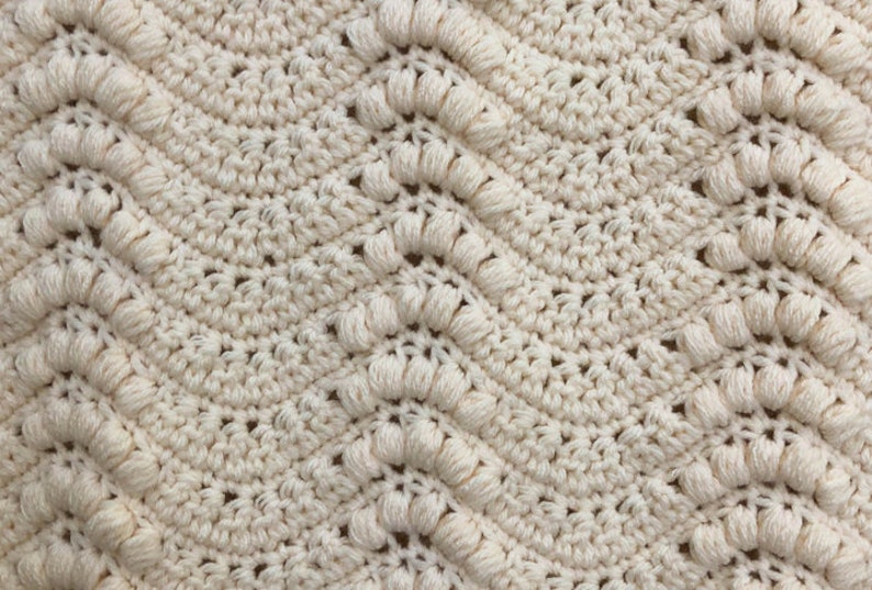 May include: A close-up of a crocheted blanket with a textured pattern. The blanket is made of a cream-colored yarn and has a repeating pattern of small, raised bumps.