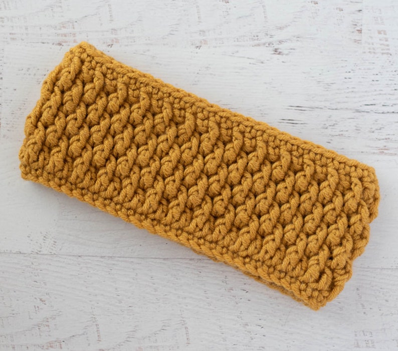 Peut inclure: Bandeau de t&ecirc;te en crochet jaune moutarde avec un motif tiss&eacute; textur&eacute;. Le bandeau est rectangulaire et semble fait &agrave; la main. Le fil a une texture l&eacute;g&egrave;rement floue. Le bandeau est pos&eacute; sur une surface en bois blanc.