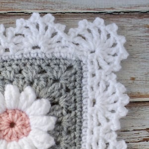 May include: A close-up of a crocheted blanket with a white crocheted border. The blanket is gray with a white and pink flower in the center.