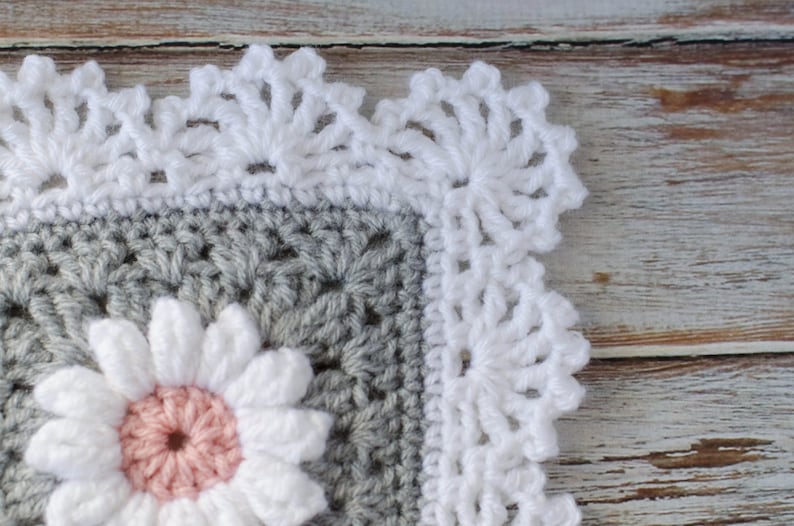 May include: A close-up of a crocheted blanket with a white crocheted border. The blanket is gray with a white and pink flower in the center.