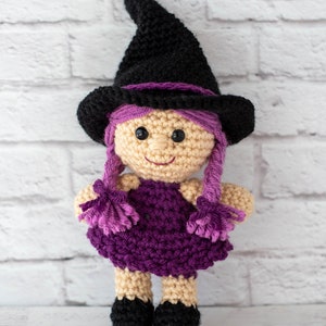 May include: A crocheted witch doll with a black pointed hat, purple hair, and a purple dress. The doll has a smiling face with black eyes and a beige complexion.