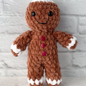 May include: A brown crocheted gingerbread man doll with white accents on the head, hands, and feet. The doll has black button eyes, a stitched smile, and red button details down the front. A handmade holiday decoration.