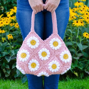 May include: A pink crocheted tote bag with white daisy flowers. The bag has a wide, flat bottom and two long straps.