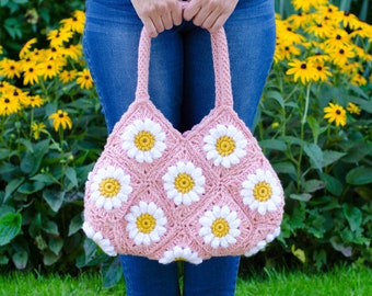May include: A pink crocheted tote bag with white daisy flowers. The bag has a wide, flat bottom and two long straps.