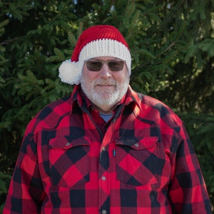 Op de afbeelding: Een man draagt een rood-witte gebreide kerstmanmuts met een witte pompon, een rood-zwart geruit overhemd en een witte baard.