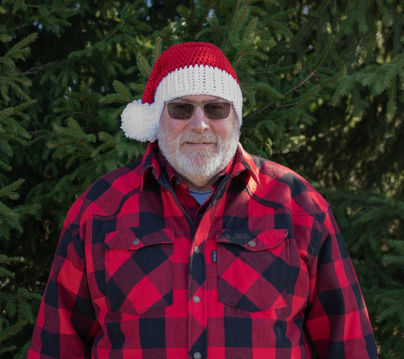 Op de afbeelding: Een man draagt een rood-witte gebreide kerstmanmuts met een witte pompon, een rood-zwart geruit overhemd en een witte baard.