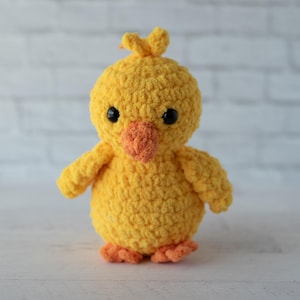 May include: A handmade, yellow crochet chick with black eyes and an orange beak and feet. The chick has small wings and a tuft of yarn on its head. The plush toy is standing on a white surface.