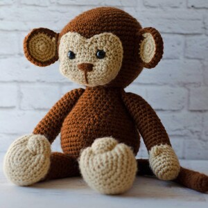 May include: A brown crocheted monkey toy with beige accents. The monkey is sitting with its legs crossed and its tail curled behind it.