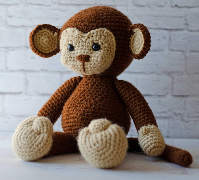 May include: A brown crocheted monkey toy with beige accents. The monkey is sitting with its legs crossed and its tail curled behind it.