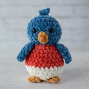 以下が含まれることがあります： 手作りのぬいぐるみ鳥のおもちゃ。青い頭と翼、赤い胸、白いお腹があります。鳥は茶色のくちばしと足、黒い目をしています。おもちゃは柔らかく、質感のある素材で作られています。