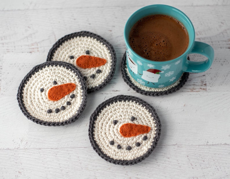 K&ouml;nnte beinhalten: Vier geh&auml;kelte Untersetzer mit Schneemann-Gesichtern. Die Untersetzer sind wei&szlig; mit grauen R&auml;ndern und haben orangefarbene Filz-Nasen und schwarze Filz-Augen. Eine blaue Tasse mit einem Schneemann-Motiv steht auf einem der Untersetzer.