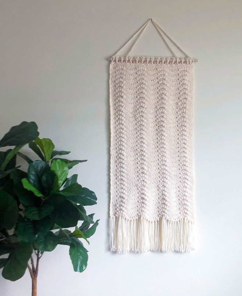May include: A white crocheted wall hanging with a fringe border. The hanging is attached to a wooden dowel rod with a white cord.