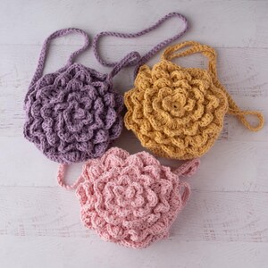 May include: Three round crochet bags with a textured, layered petal design. One bag is light purple, one is mustard yellow, and one is light pink. Each bag features a matching crocheted strap attached to the top, creating a floral appearance.