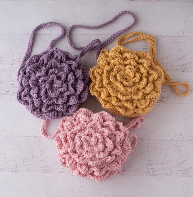 May include: Three round crochet bags with a textured, layered petal design. One bag is light purple, one is mustard yellow, and one is light pink. Each bag features a matching crocheted strap attached to the top, creating a floral appearance.