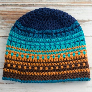 May include: A handmade crochet beanie in shades of navy blue, teal, brown, and gold. The hat features a striped pattern with textured stitches. The beanie is designed to keep the wearer warm and stylish during colder weather.