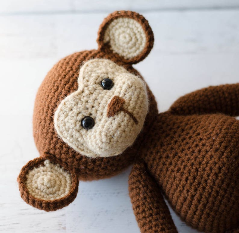 May include: A brown crocheted monkey toy with beige ears and a beige face. The monkey has black eyes and a small brown nose.