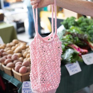 May include: A light pink reusable market bag crafted from crocheted yarn. The bag features an open, diamond-shaped mesh pattern throughout the body, a solid crocheted base, and two long, thin crocheted handles for carrying items by hand.