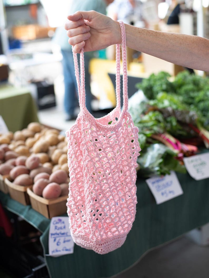 May include: A light pink reusable market bag crafted from crocheted yarn. The bag features an open, diamond-shaped mesh pattern throughout the body, a solid crocheted base, and two long, thin crocheted handles for carrying items by hand.
