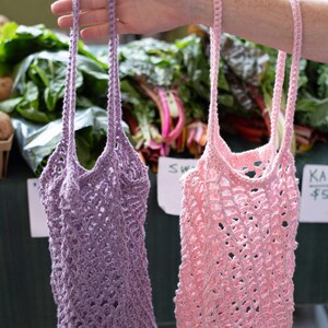 May include: Two reusable market bags made of crocheted yarn, one in a light lavender purple and the other in a soft pastel pink. Each bag features an open-weave mesh pattern, a circular base, and two long, slender handles for carrying by hand or shoulder.