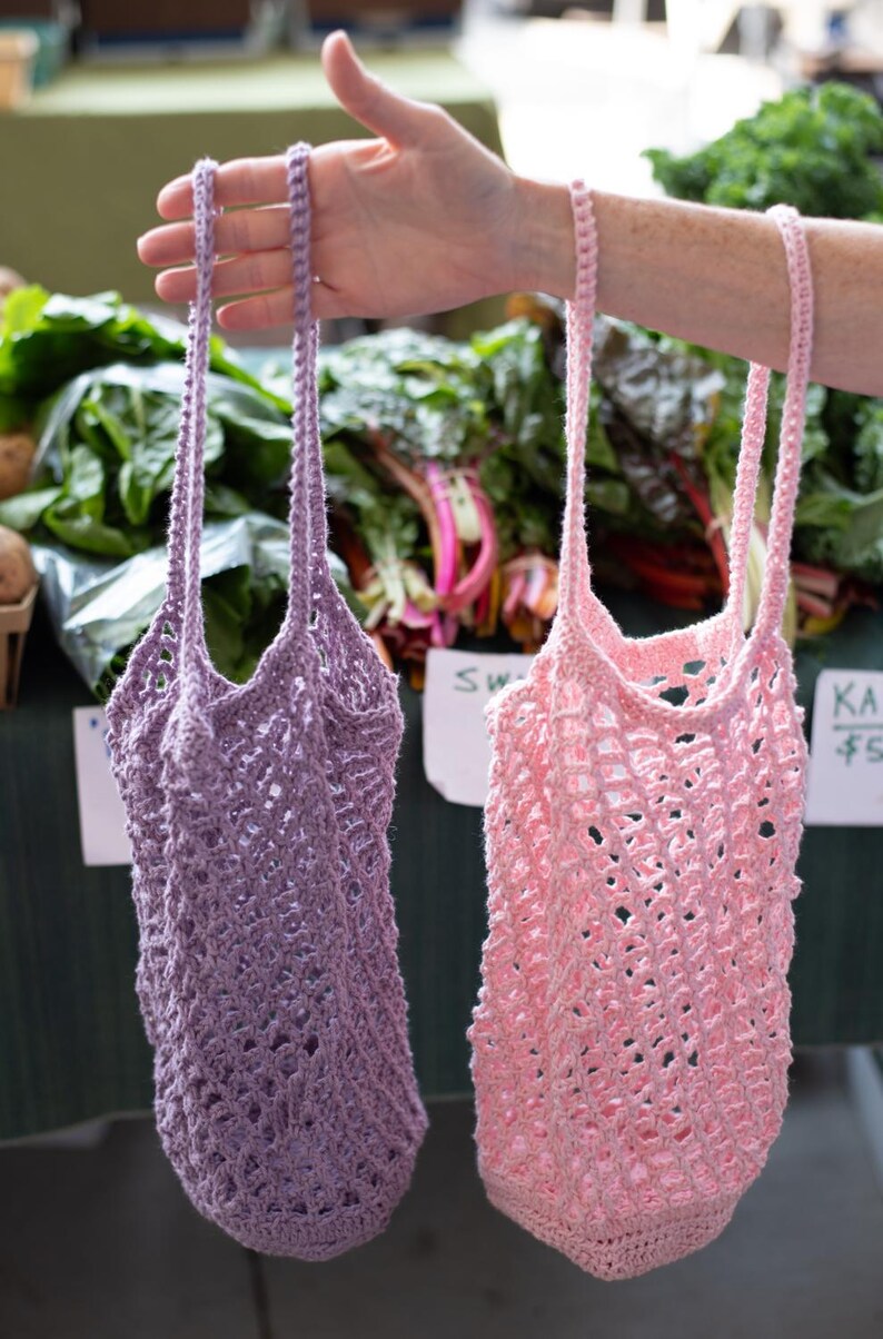 May include: Two reusable market bags made of crocheted yarn, one in a light lavender purple and the other in a soft pastel pink. Each bag features an open-weave mesh pattern, a circular base, and two long, slender handles for carrying by hand or shoulder.
