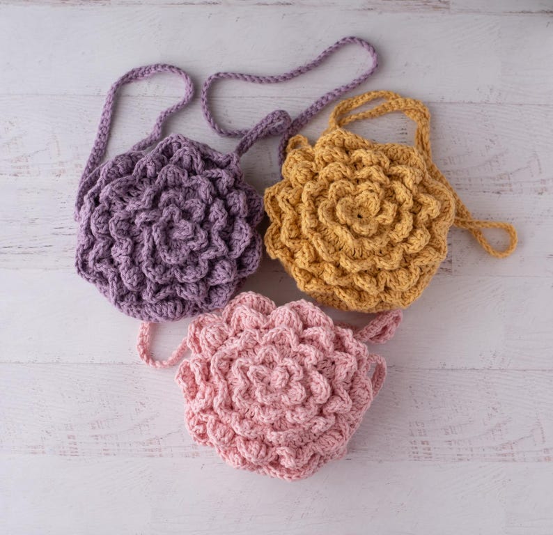May include: Three round crochet bags with a textured floral petal design. One bag is purple, one is mustard yellow, and one is light pink. Each bag features a matching crocheted shoulder strap and a layered, dimensional surface pattern.