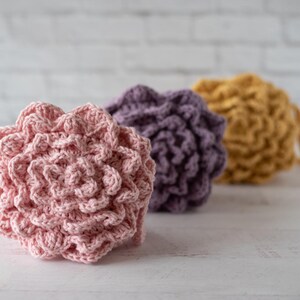 May include: Three round, textured crochet scrubbers arranged in a row. The front scrubber is light pink, the middle one is purple, and the back one is mustard yellow. Each item features a dense, layered, floral-like pattern made of yarn.