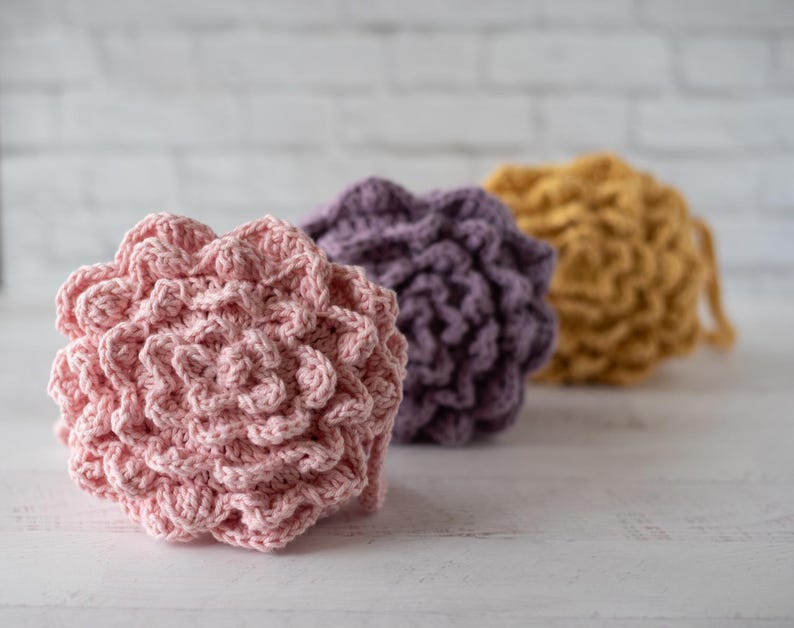 May include: Three round, textured crochet scrubbers arranged in a row. The front scrubber is light pink, the middle one is purple, and the back one is mustard yellow. Each item features a dense, layered, floral-like pattern made of yarn.