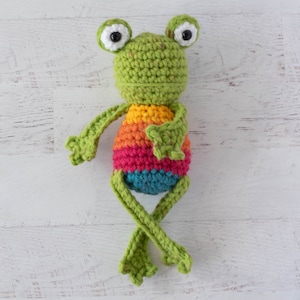 Puede incluir: Un juguete de rana de crochet con cuerpo verde, rayas de colores arcoíris y ojos negros.