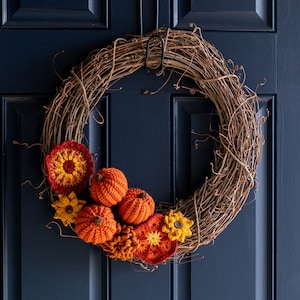 May include: A grapevine wreath decorated with crocheted pumpkins and flowers in shades of orange, yellow, and red, perfect for autumn.