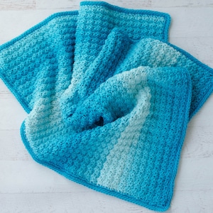 May include: A crocheted baby blanket in shades of blue, with a textured stitch pattern. The blanket is folded in half and has a light blue border.