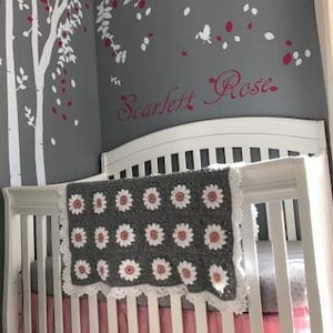May include: A white crib with a gray and pink crocheted blanket with a white lace trim. The crib has a pink ruffle around the bottom. The wall behind the crib is gray with white and pink floral decals and the name "Scarlett Rose" written in pink.
