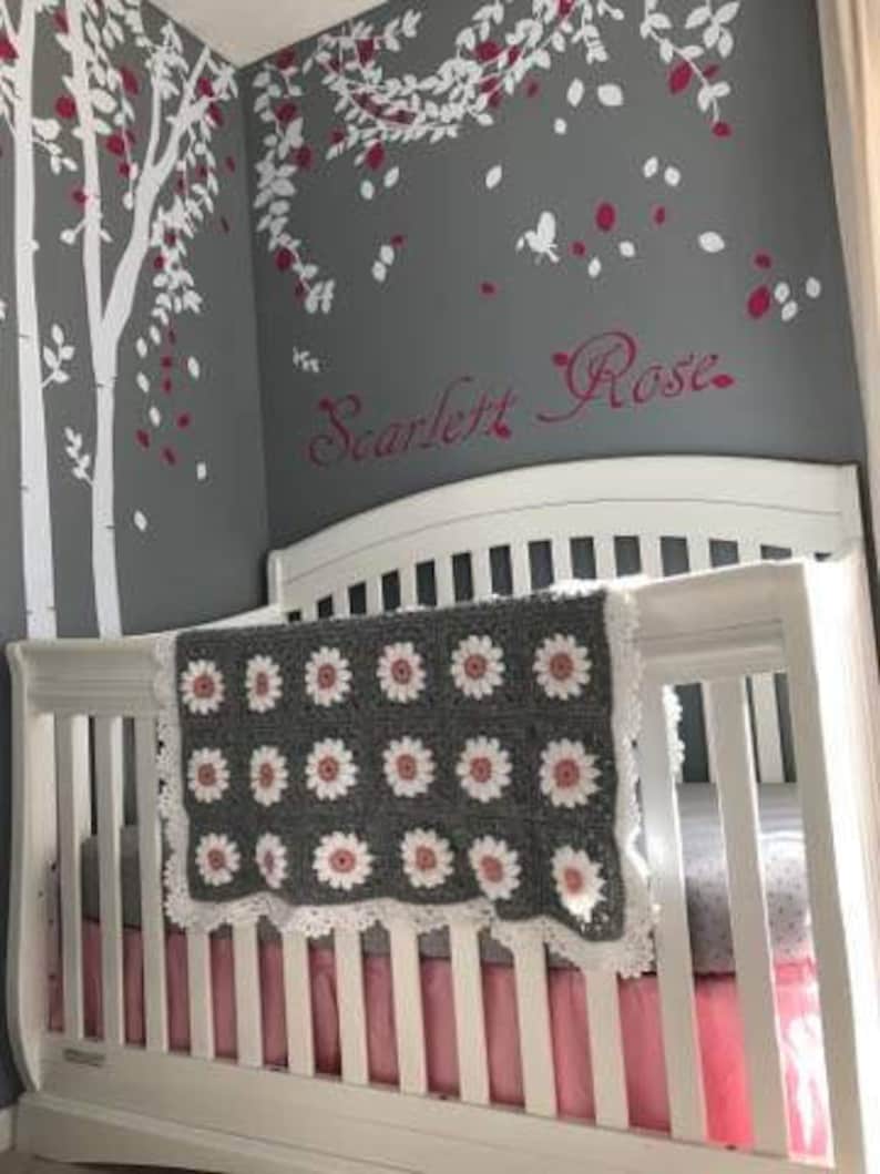 May include: A white crib with a gray and pink crocheted blanket with a white lace trim. The crib has a pink ruffle around the bottom. The wall behind the crib is gray with white and pink floral decals and the name "Scarlett Rose" written in pink.
