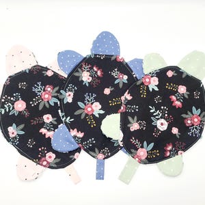 May include: Three handmade turtle-shaped baby toys. Each turtle has a black body with a floral pattern in pink, white, and green. The flippers and head are in various pastel colors with polka dots. The toys are made of soft fabric.